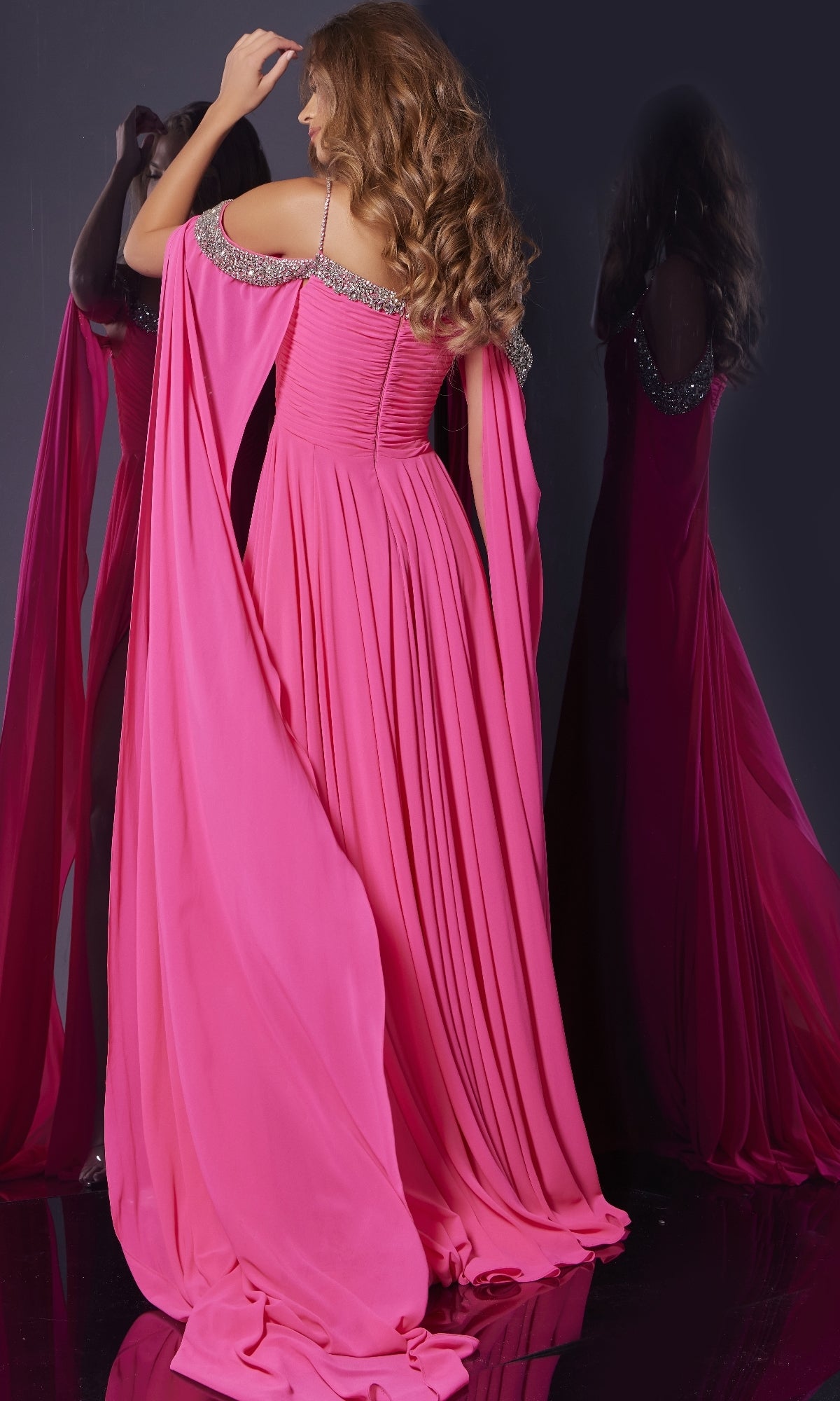 A woman wearing the Long Formal Dress: Jovani 38996—an off-the-shoulder pink gown with silver embellishments—faces a dark mirror, her wavy hair cascading down her back.
